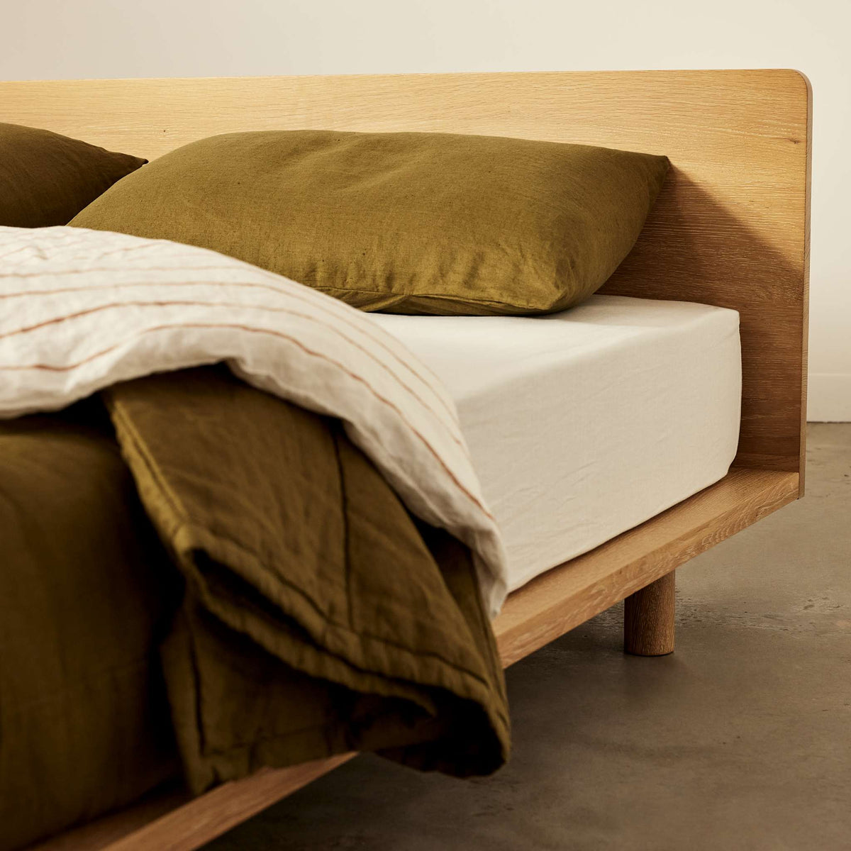 Corner view of Lagos timber bed frame in solid oak, styled with olive green linen and white sheet set - Totem Road